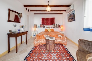 A bedroom with a bed, a wooden nightstand, a bedside table, a chair, a patterned rug, a hanging light fixture, and a wall-mounted air conditioner.