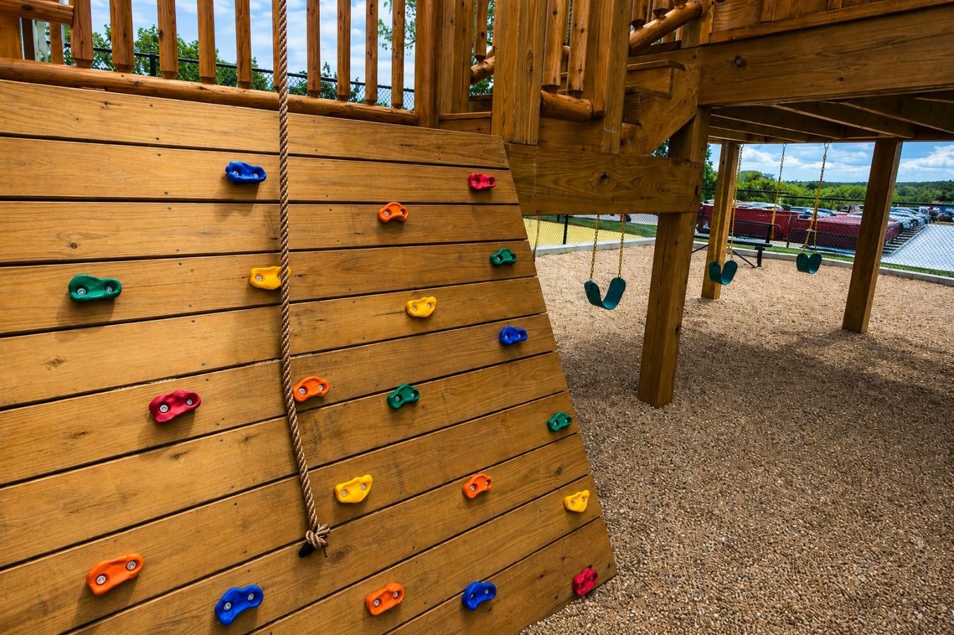 Una pared de escalar de madera con presas de distintos colores, una cuerda y un conjunto de columpios al fondo.
