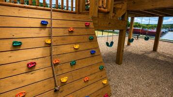 Una pared de escalar de madera con presas de distintos colores, una cuerda y un conjunto de columpios al fondo.