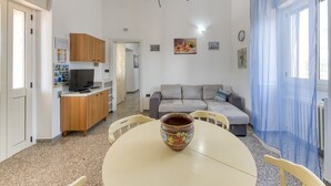 A living room with a sofa, a dining table with a decorative pot, a kitchen area with cabinets, and a blue curtain. - Holiday Home 'Villetta Il Boschetto' with Shared Pool, Private Garden and Air Conditioning (Morciano di Leuca)