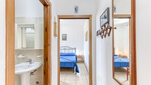 Un pasillo estrecho con un baño a la izquierda, una cama con colcha azul en el centro y un espejo a la derecha.
