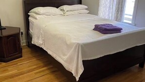 Een netjes opgemaakt bed met witte lakens en een paarse handdoek, houten vloeren en een nachtkastje met een lamp.