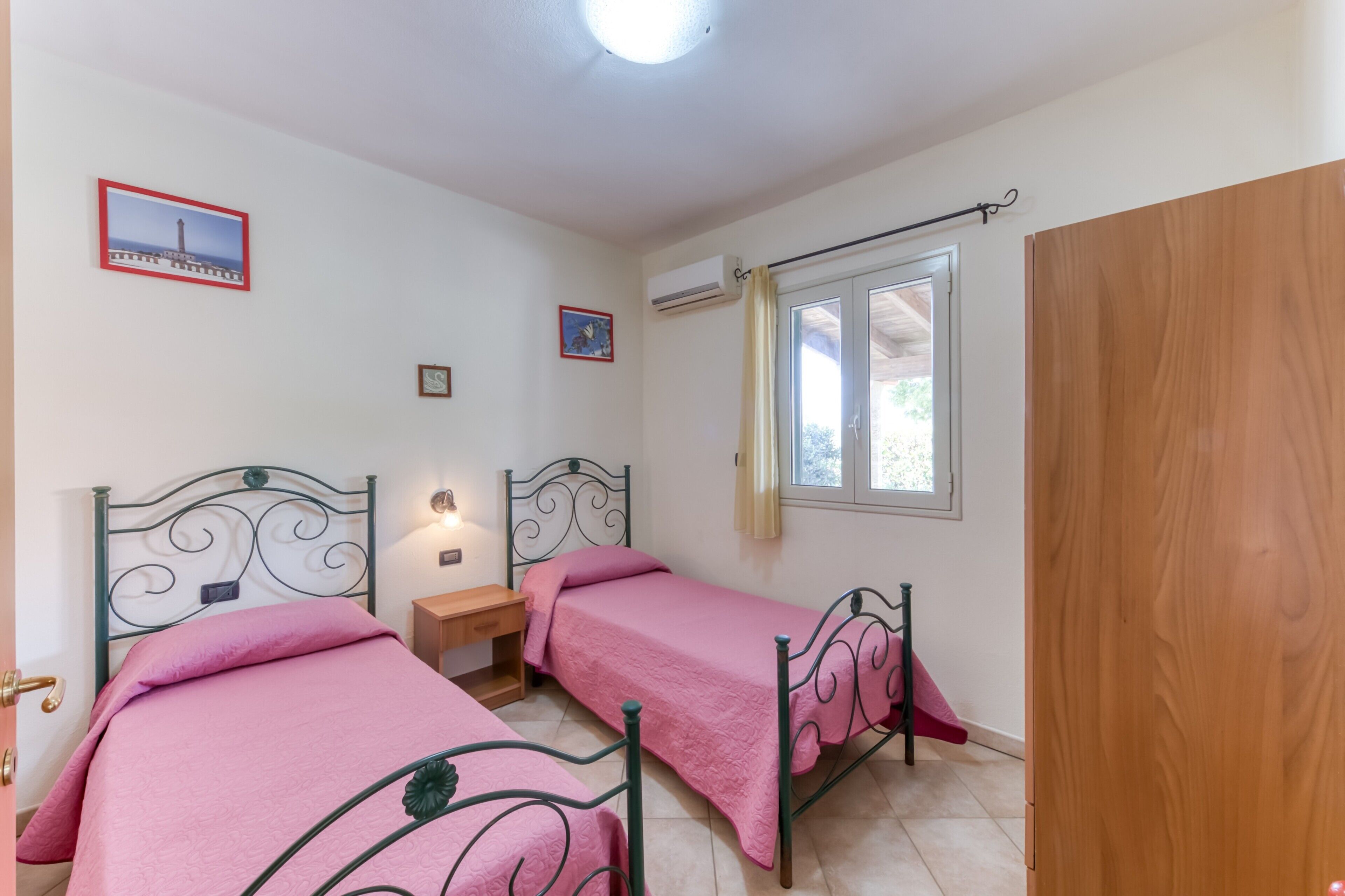 A bedroom with two beds, a wooden door, a window with a curtain, and a wall-mounted air conditioner.