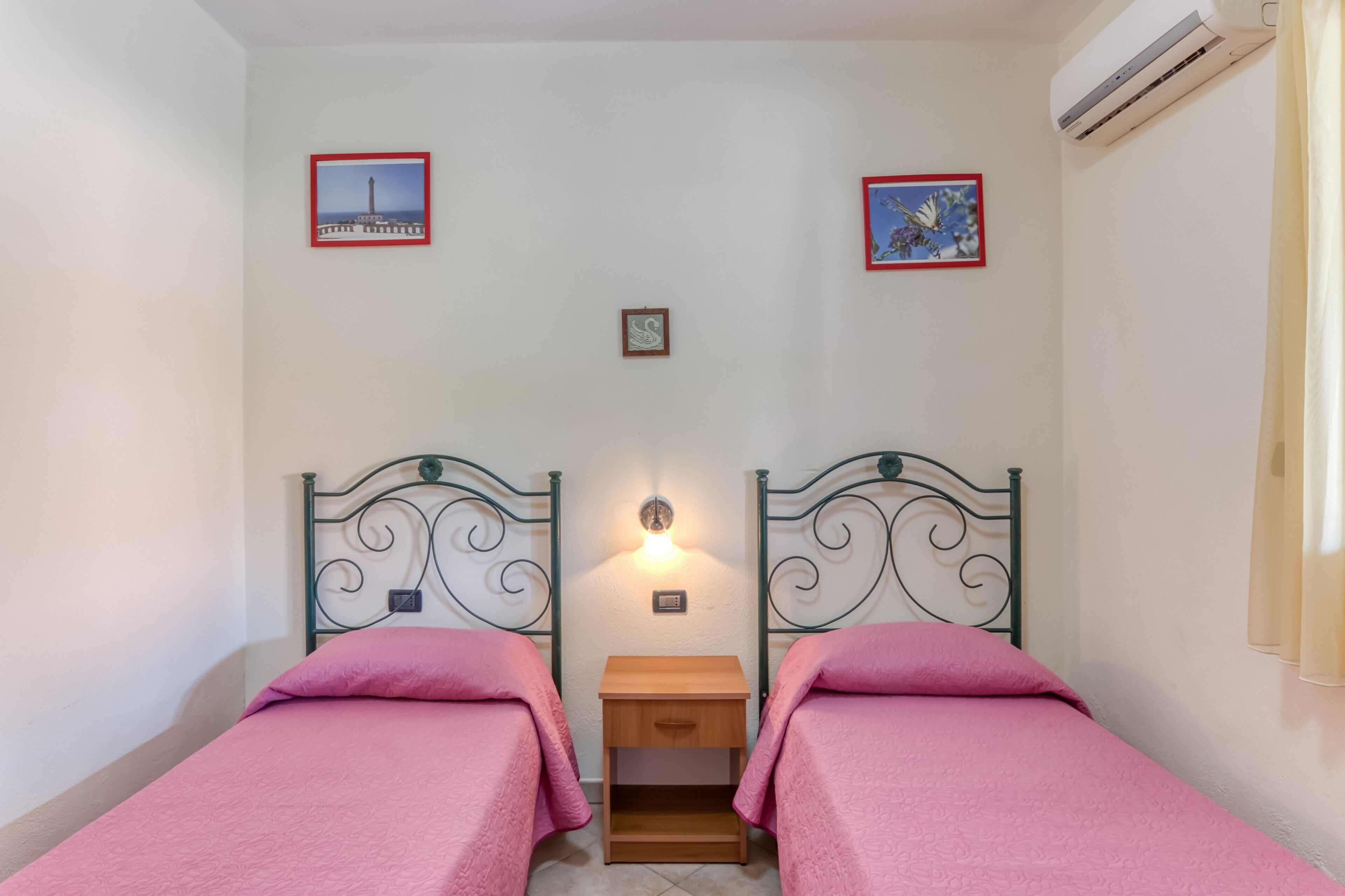 Habitació amb dos llits, una tauleta de nit, una làmpada penjada a la paret i dos quadres amb marc.