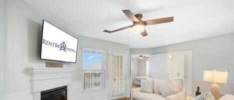 A living room with a fireplace, a ceiling fan, a television mounted on the wall, and a couch with patterned cushions.