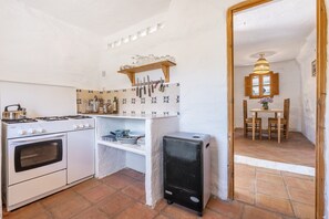 Een compacte keuken met een gasfornuis, oven en een kleine koelkast. Een plank met keukengerei en een eethoek met een tafel en stoelen.