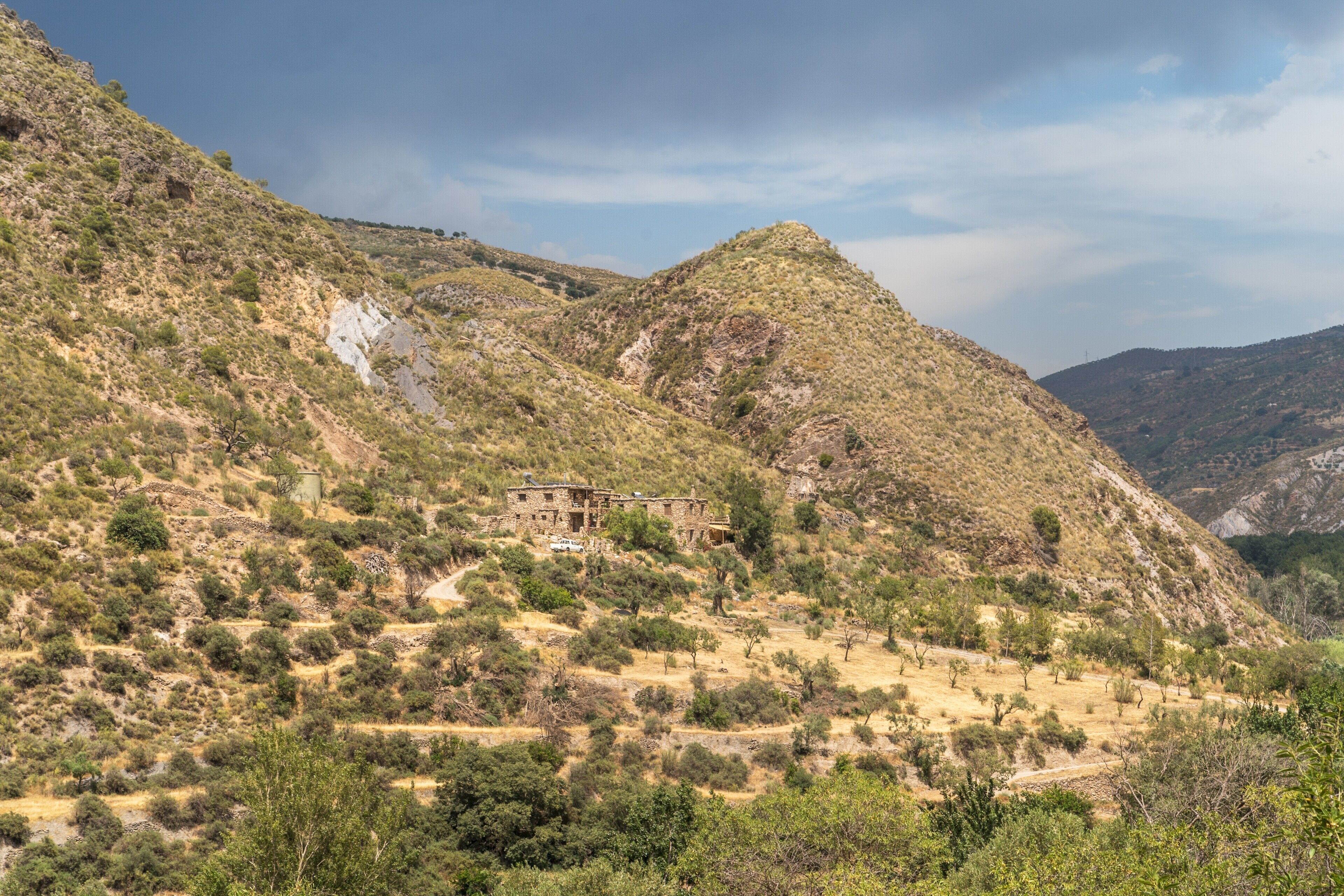 Un paisaje montañoso con una construcción rústica de piedra rodeada de vegetación seca y un cielo despejado.