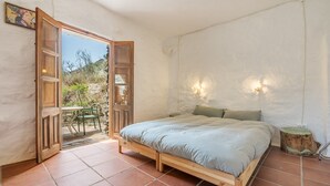 A bedroom with a wooden bed, a small table, two wall-mounted lights, and a view of the outdoors.