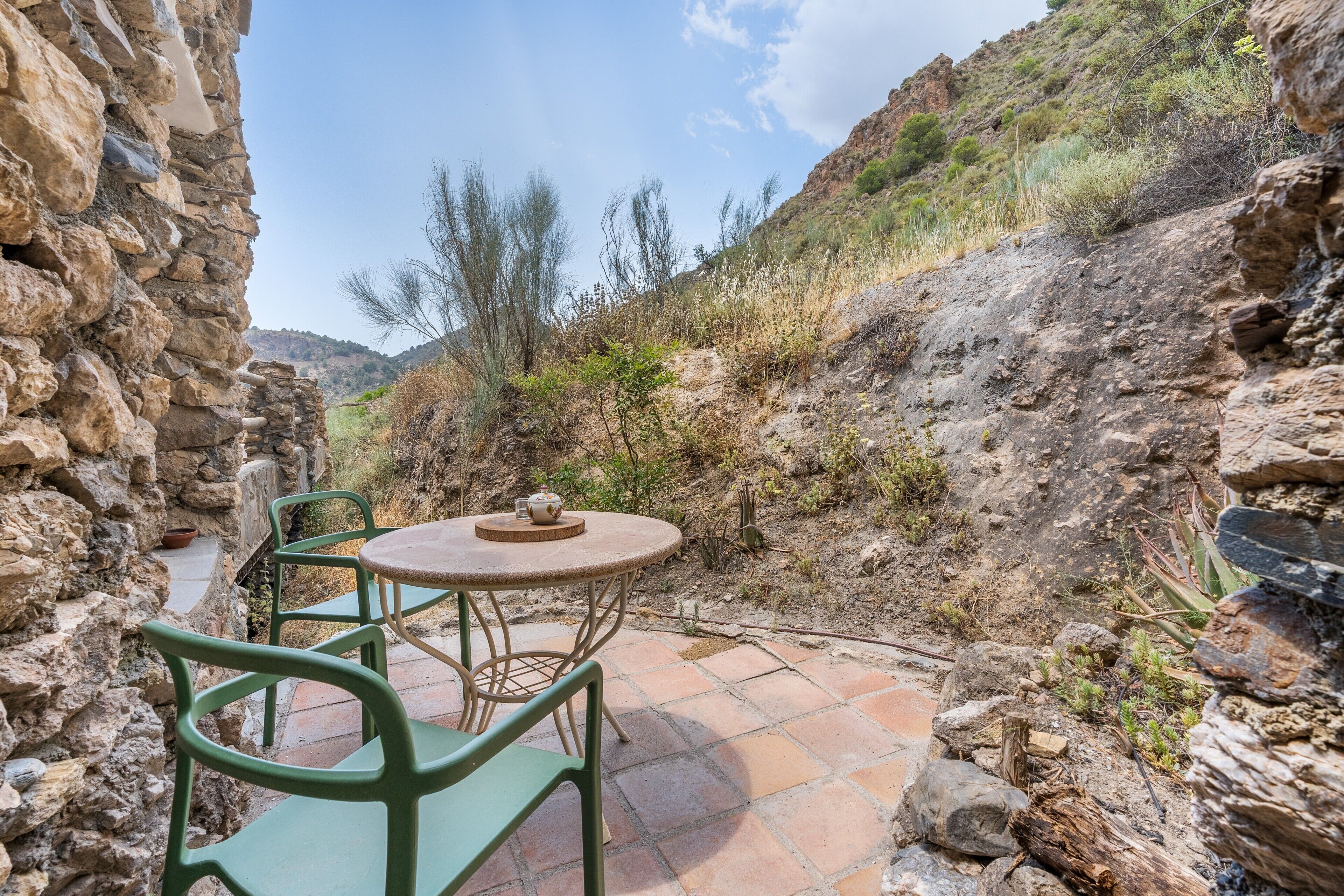 Un espace extérieur aménagé avec des murs en pierre, comprenant une table et des chaises, et entouré d’un paysage naturel et de végétation.