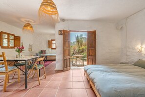 Una habitació amb un llit, una taula de dinar amb cadires, una taula petita amb una cadira i una vista a l'exterior per una porta oberta.