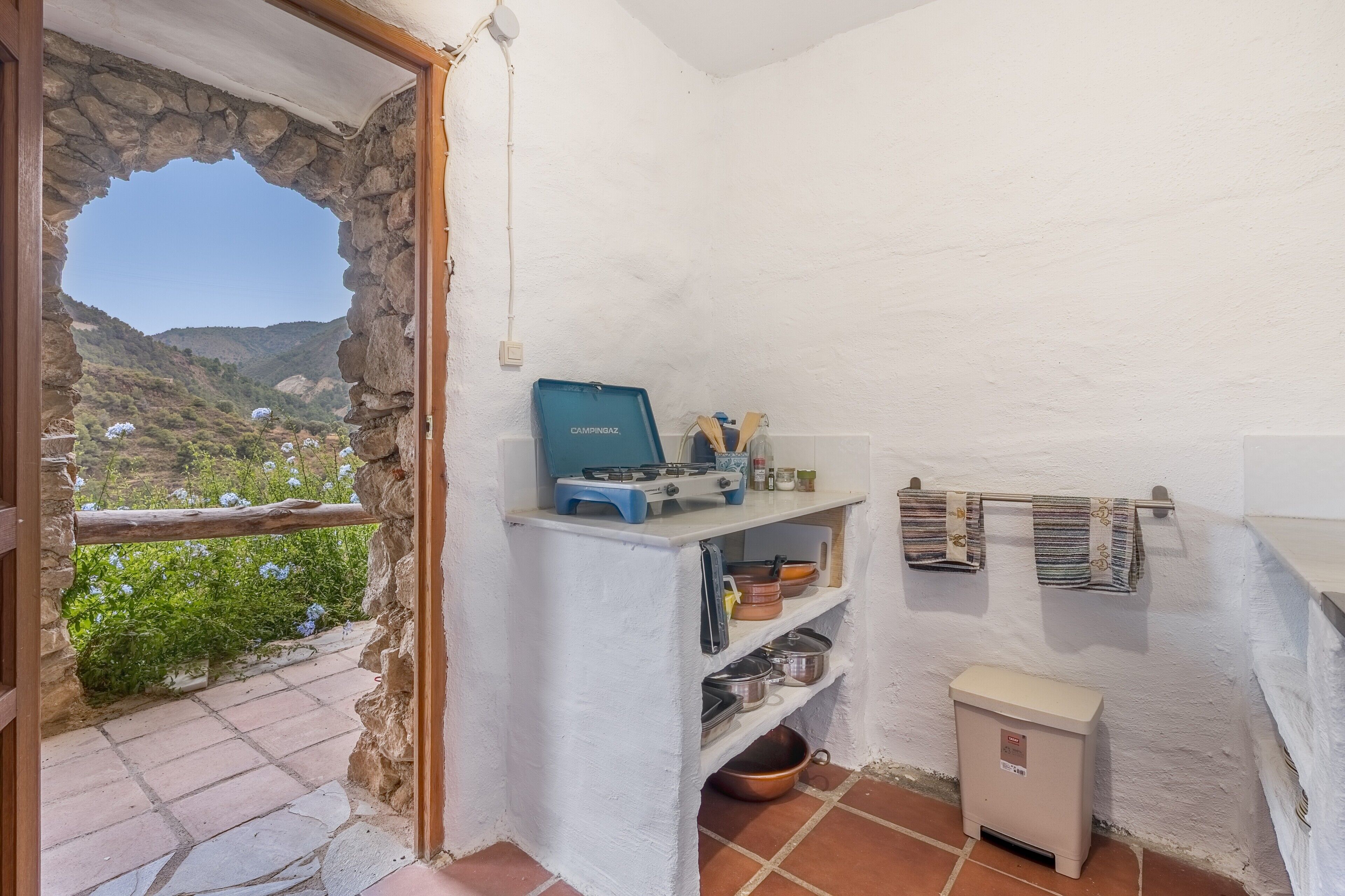 Una pequeña cocina con un ventanal de arco de piedra, una estufa de camping azul y una pared blanca con un estante que contiene utensilios de cocina.