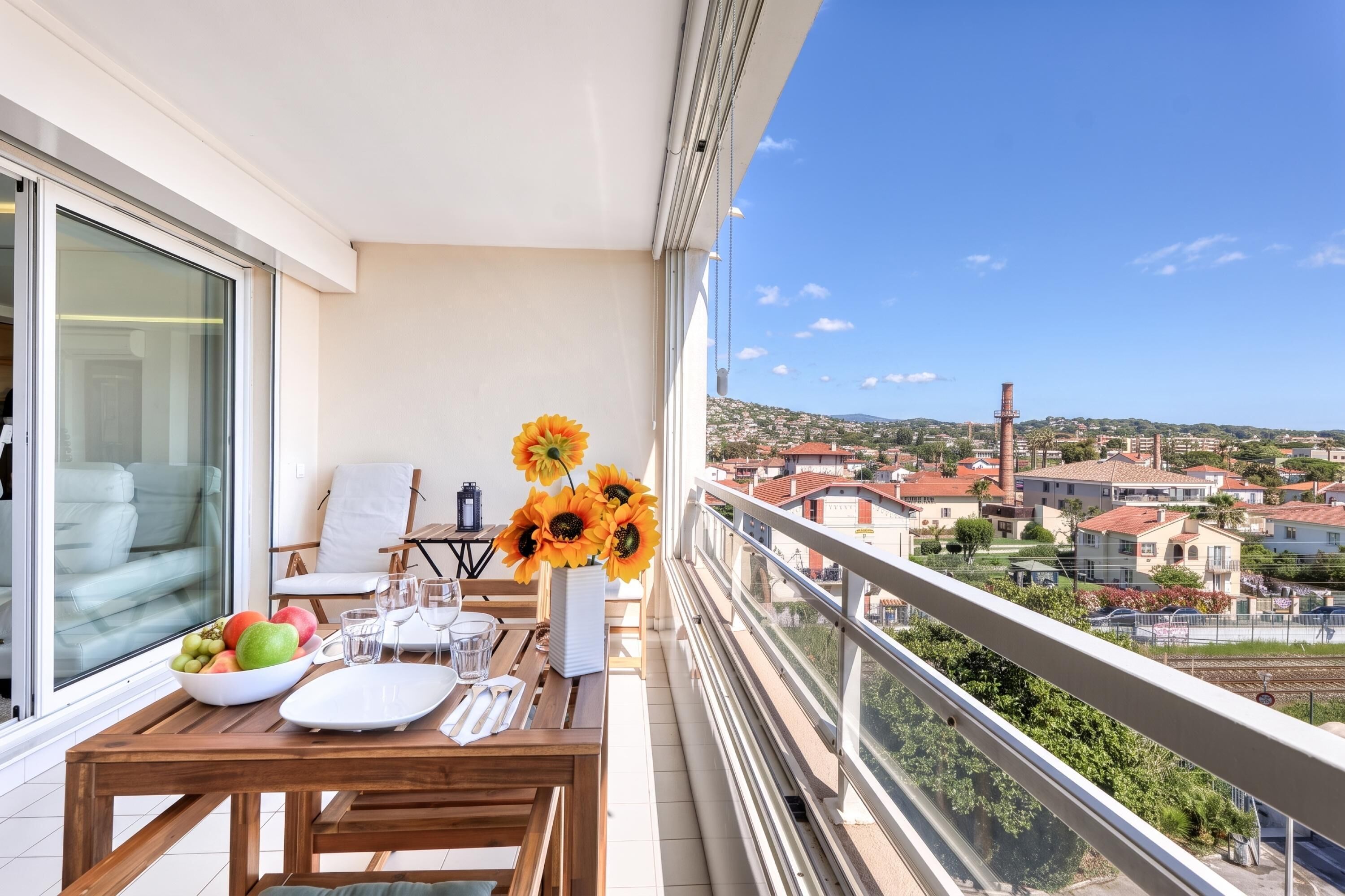 A balcony with a dining table set for two, a chair, a vase with sunflowers, and a view of a town with buildings and a tower.