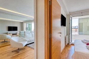 A modern hotel room with a wooden door, a flat-screen TV, a sofa, a chair, and a bed.