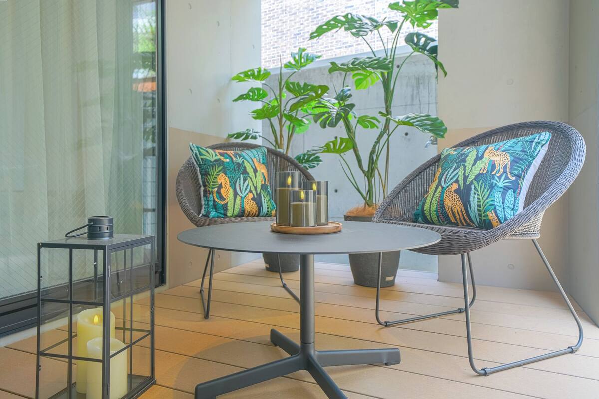 A small, round table with two wicker chairs, each with a colorful cushion. A candle holder with a candle is on the table. There are large green plants in the background.