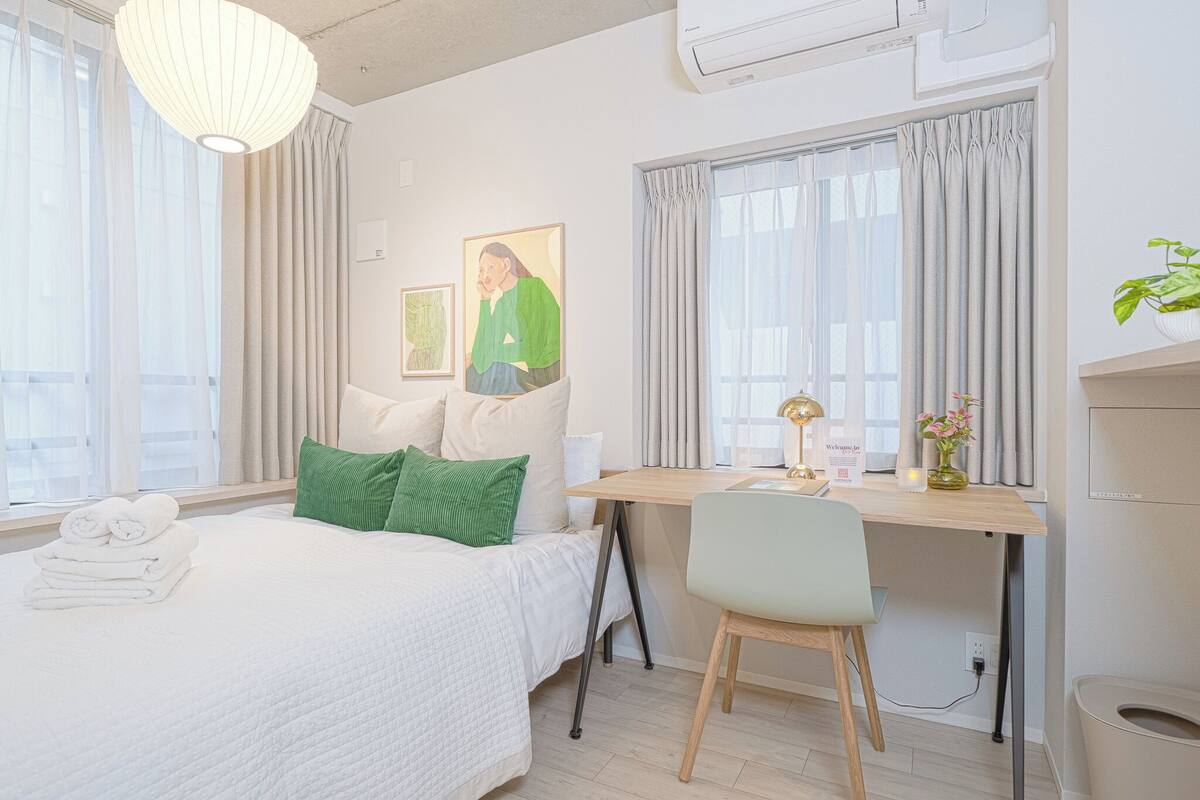A hotel room with a bed, a desk, a chair, a window with curtains, a wall-mounted air conditioner, and a welcome sign.