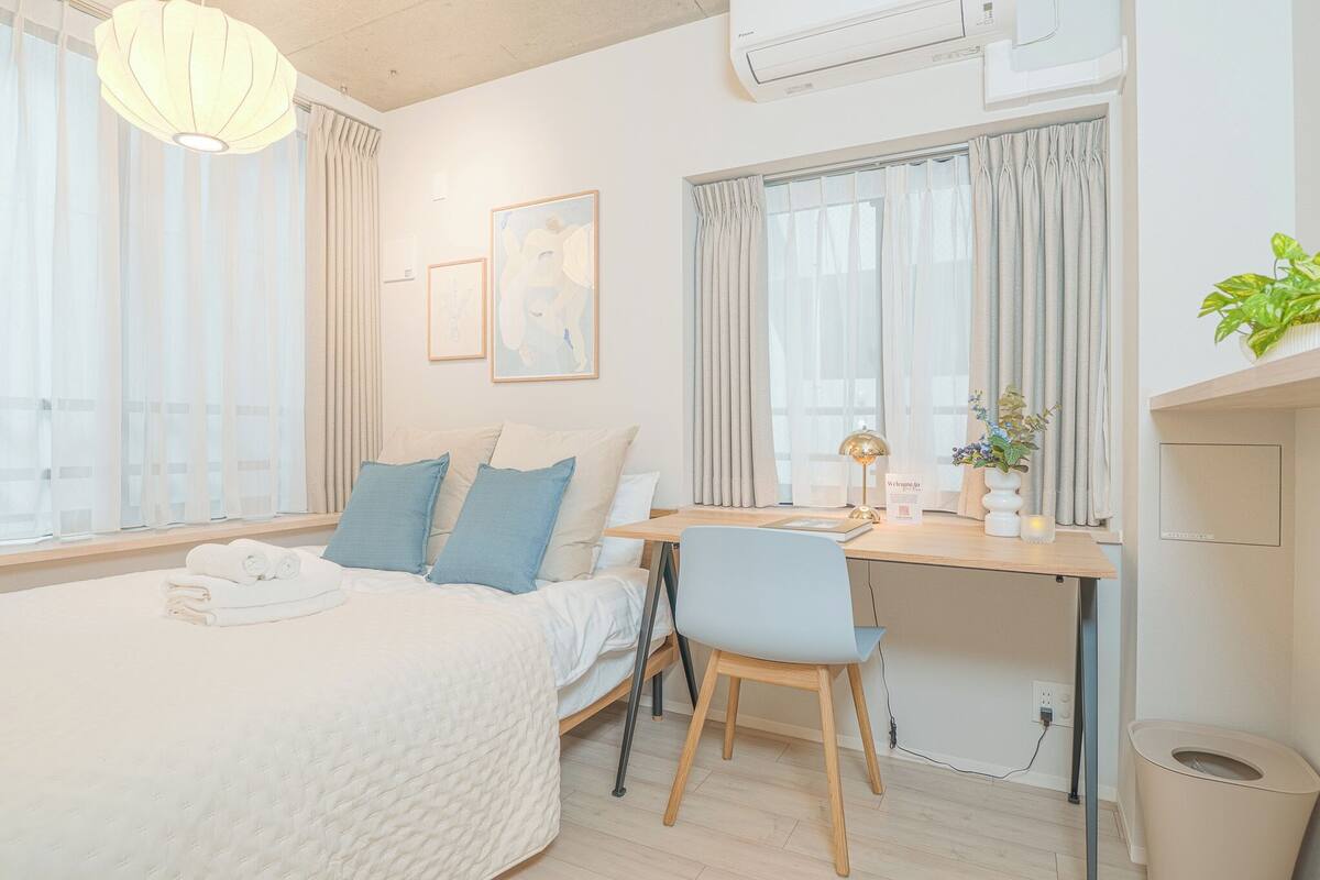 A modern hotel room with a bed, a desk, and a chair. There are curtains on the windows, a plant on the desk, and a trash bin.