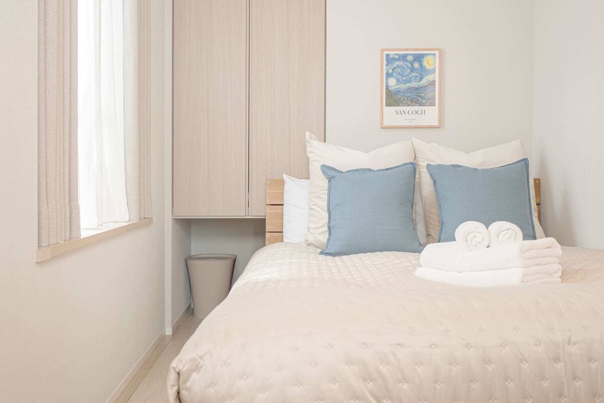 A neatly made bed with a quilted cover, a framed artwork on the wall, and a window with curtains.