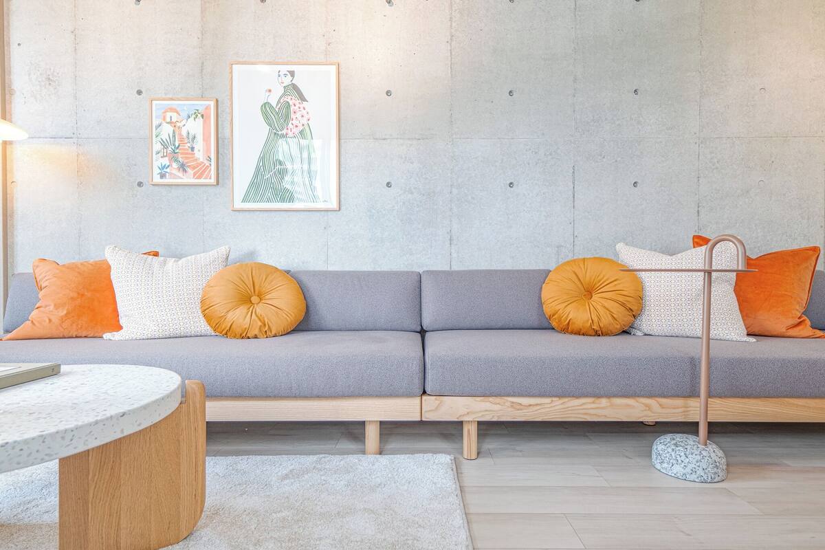 A modern living room with a grey sofa, orange and white cushions, a round wooden side table, and two framed artworks on the wall.
