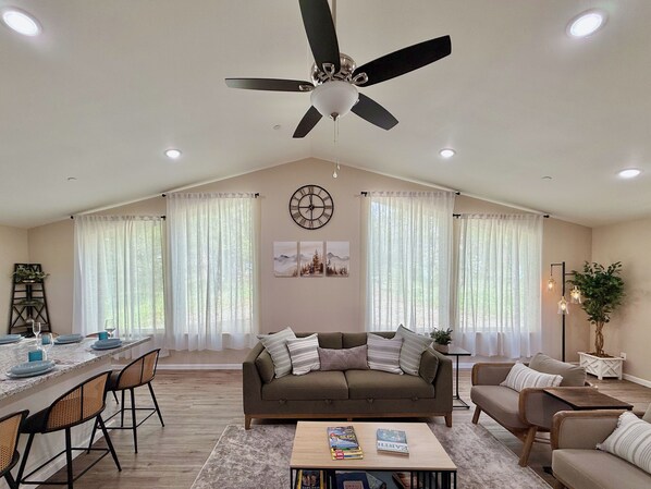 A spacious living room with a high ceiling, a ceiling fan, a dining area with a table set for four, and a large window with sheer curtains.