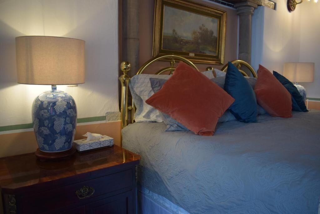 A neatly made bed with a blue and white patterned vase on a wooden nightstand, a painting on the wall, and a lamp on the left.