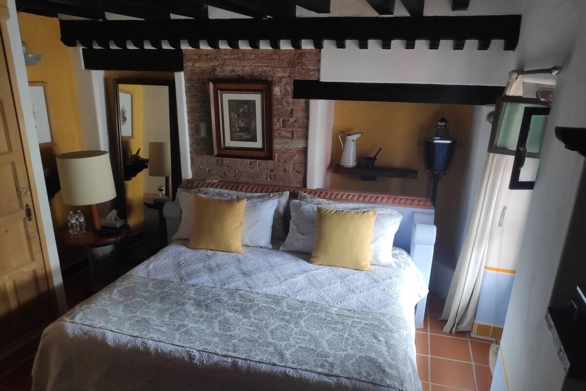 A bedroom with a bed, two pillows, a nightstand, a lamp, a picture frame, a brick wall, and a window.
