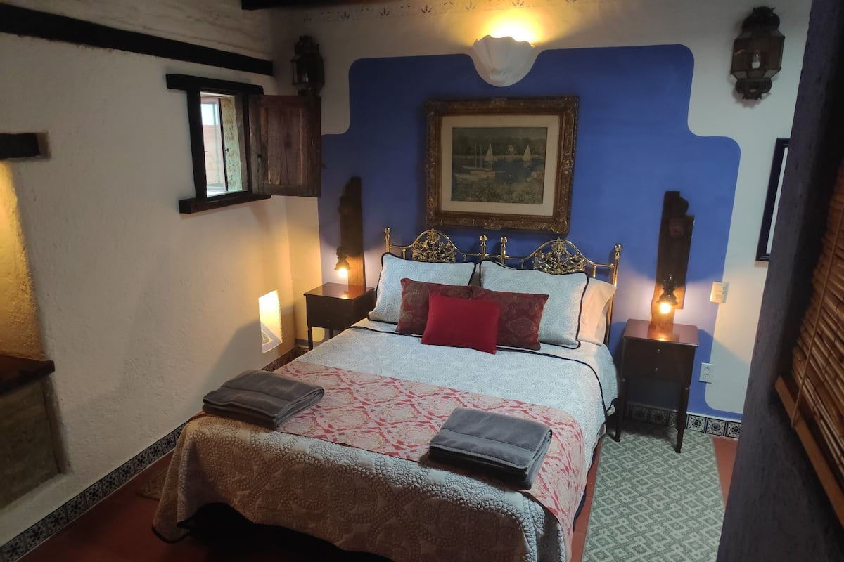 A bedroom with a bed, two bedside tables with lamps, a nightstand, a window, and a wall-mounted picture.