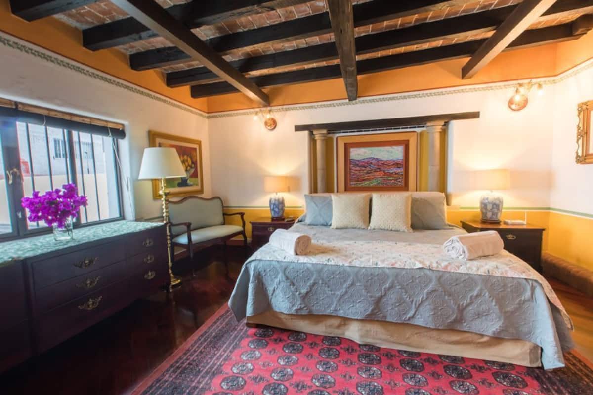 A bedroom with a large bed, a nightstand, a dresser, a chair, and a window with a view.