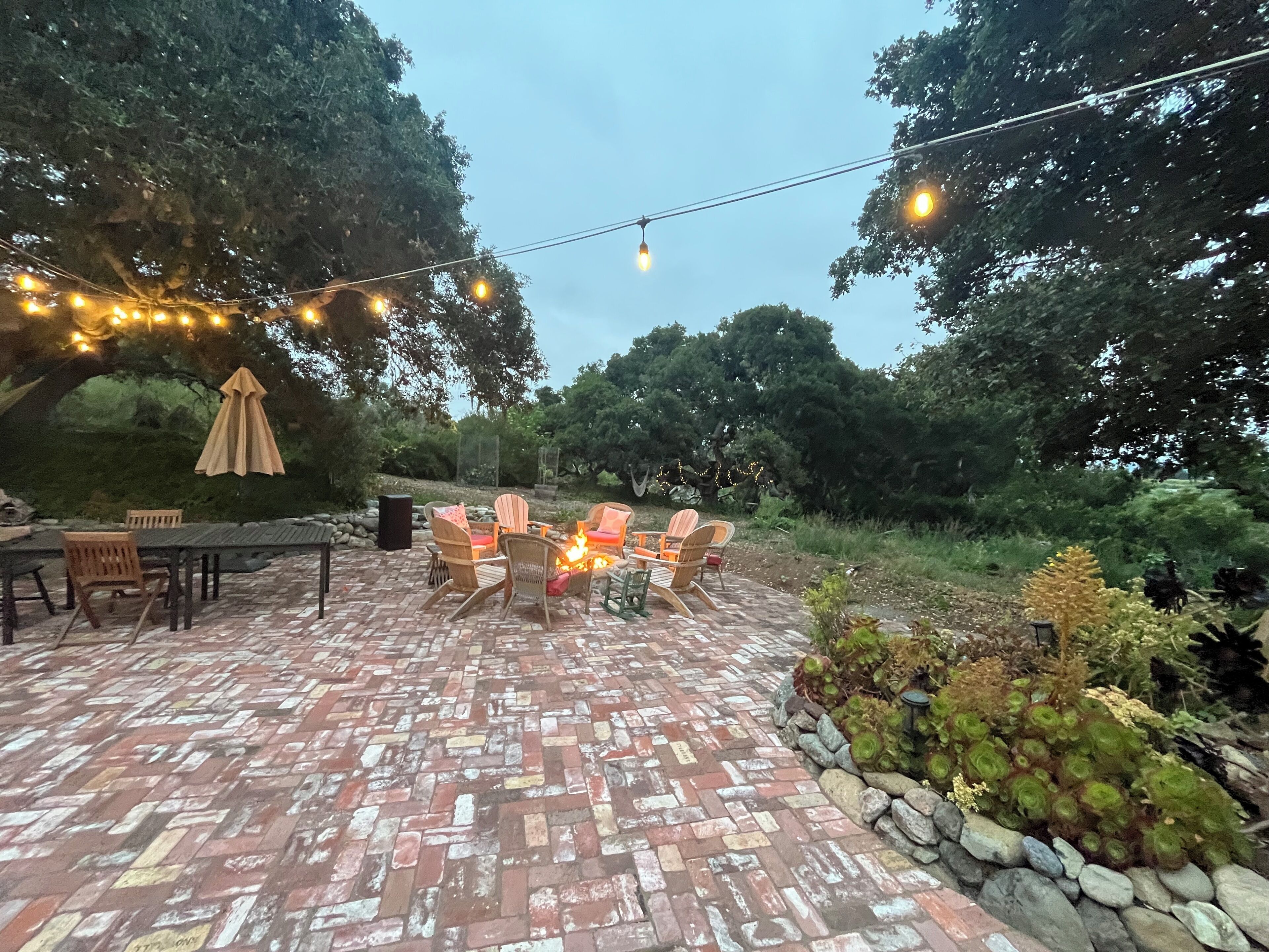 Eine Terrasse im Freien mit Backsteinboden, Gartenmöbeln und Lichterketten.