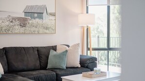 A modern living room with a dark grey sofa, a small white table, and a lamp. A framed picture of a rural scene hangs on the wall. - Blackwood at Boardwalk Villas Metung (Metung)
