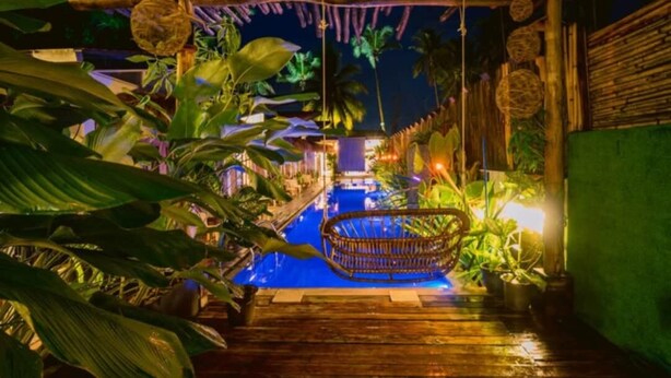 Zona piscina notturna con terrazza in legno, una sedia a dondolo e piante tropicali.