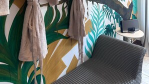 Una stanza con un grande murale tropicale, due accappatoi appesi a un appendiabiti, un divano in vimini e un tavolino con una lampada.
