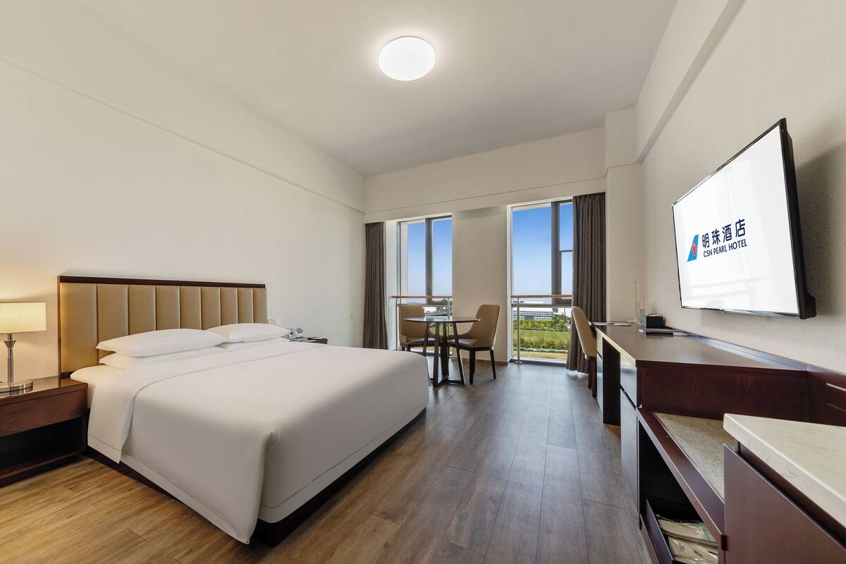 A hotel room with a bed, bedside table, lamp, TV, and a balcony with a view.