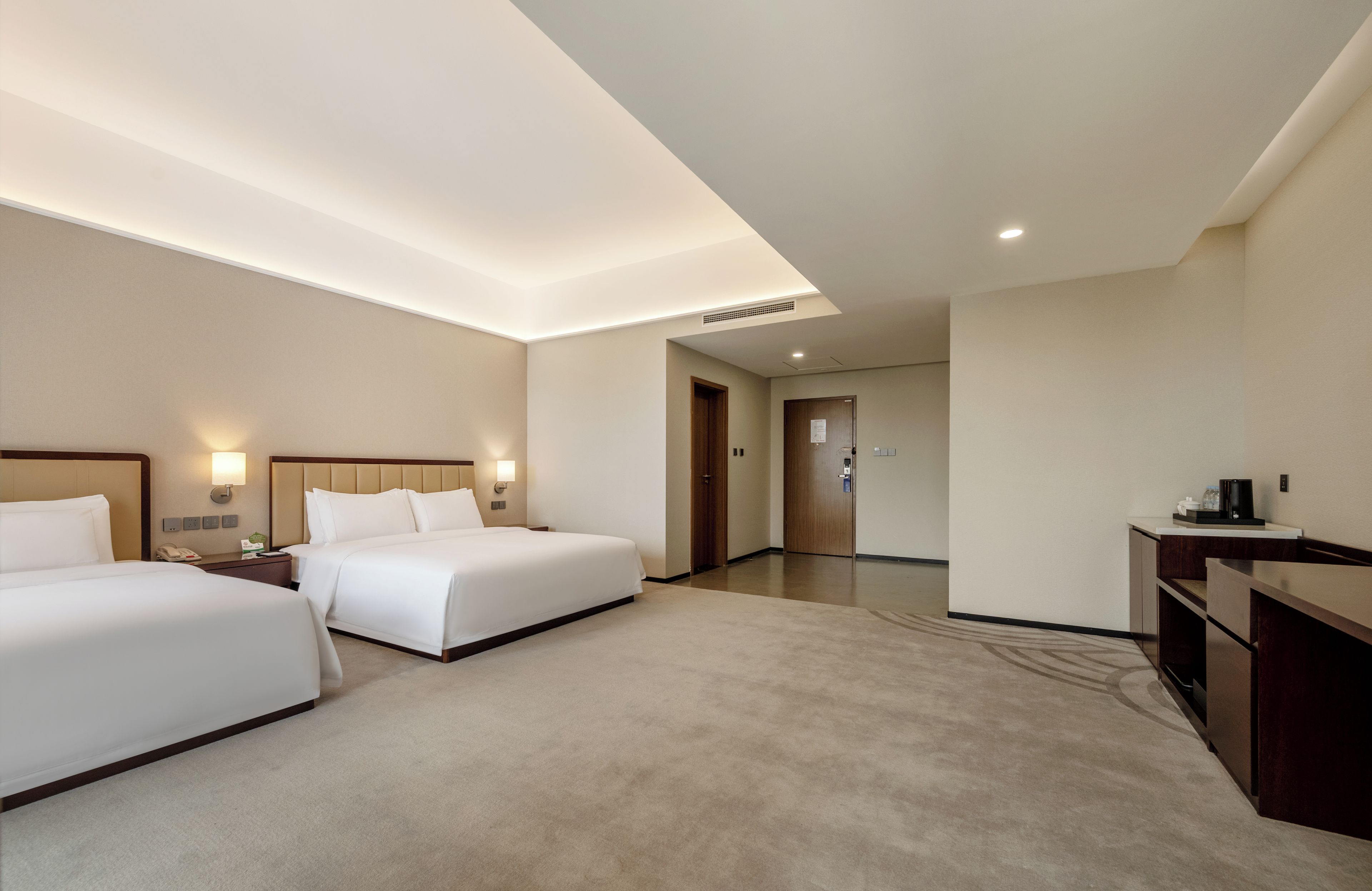 A hotel room with two beds, a desk, a coffee maker, and a door leading to a hallway.