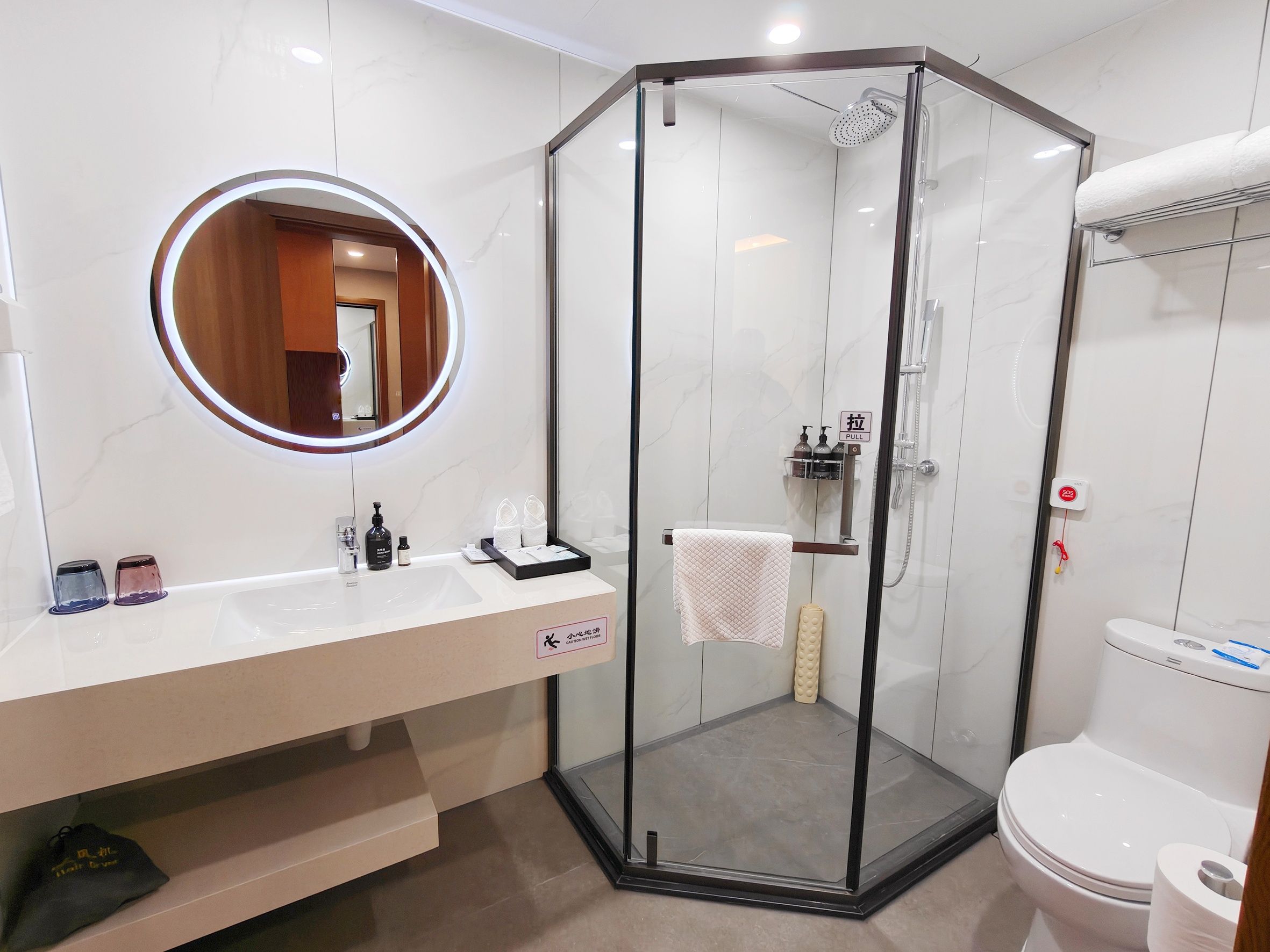 Un baño moderno con ducha envuelta en cristal, un espejo redondo, un lavabo blanco con artículos de aseo y un inodoro.