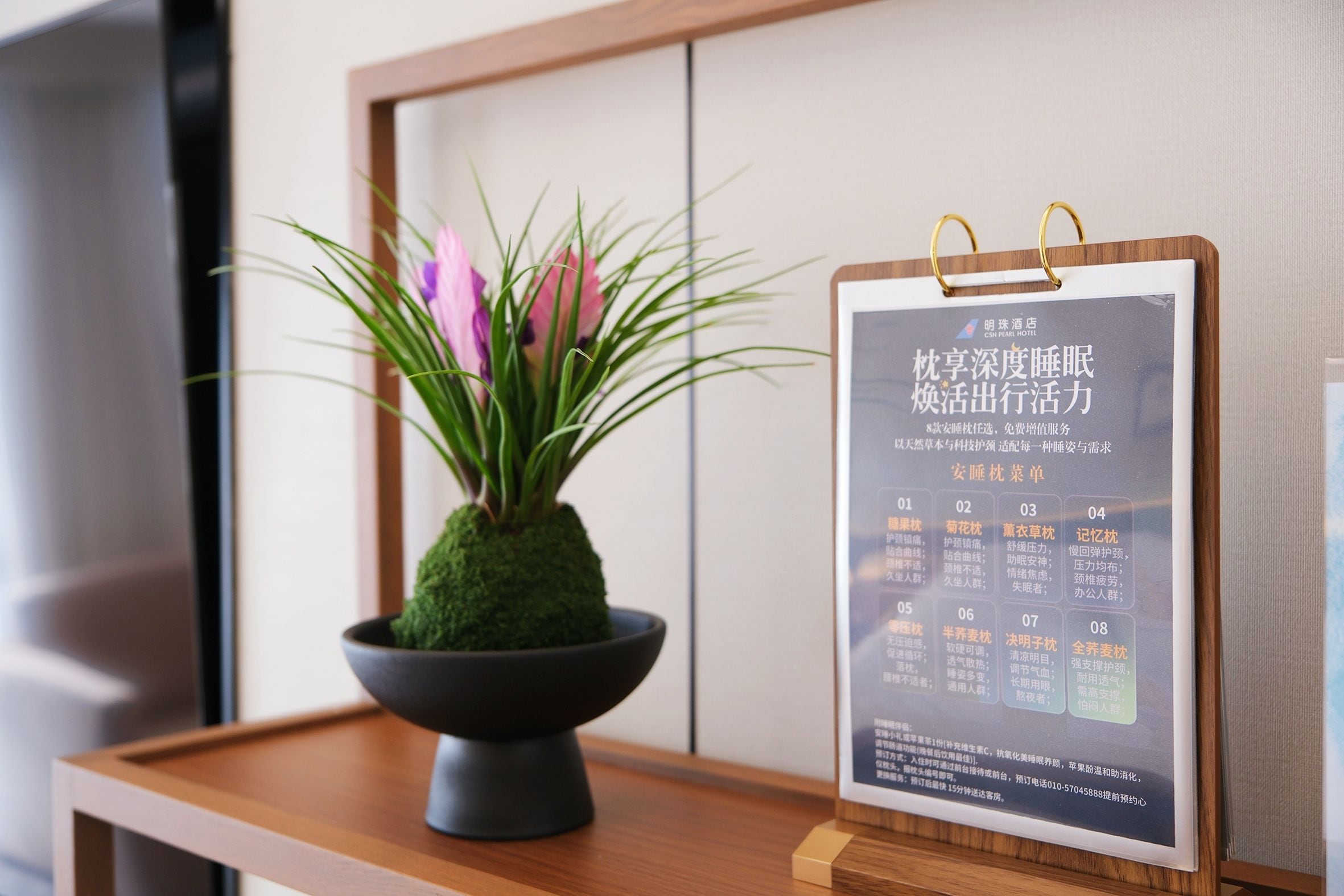 Ein Holztisch mit einer schwarzen Schüssel, in der sich eine grüne Pflanze und rosa Blumen befinden. An dem Tisch ist eine gerahmte Speisekarte oder eine Infotafel befestigt.