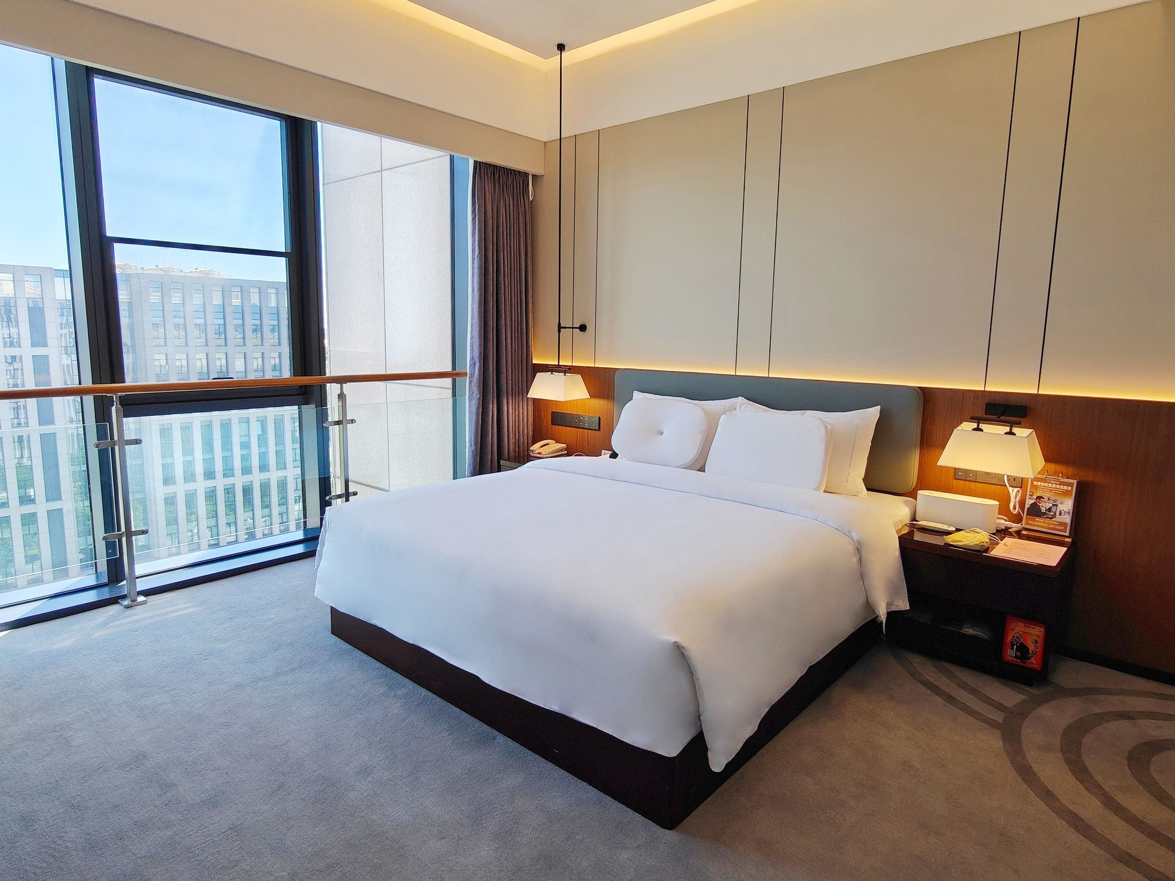 A hotel room with a large bed, a nightstand with a lamp, a telephone, and a view of buildings through the window.