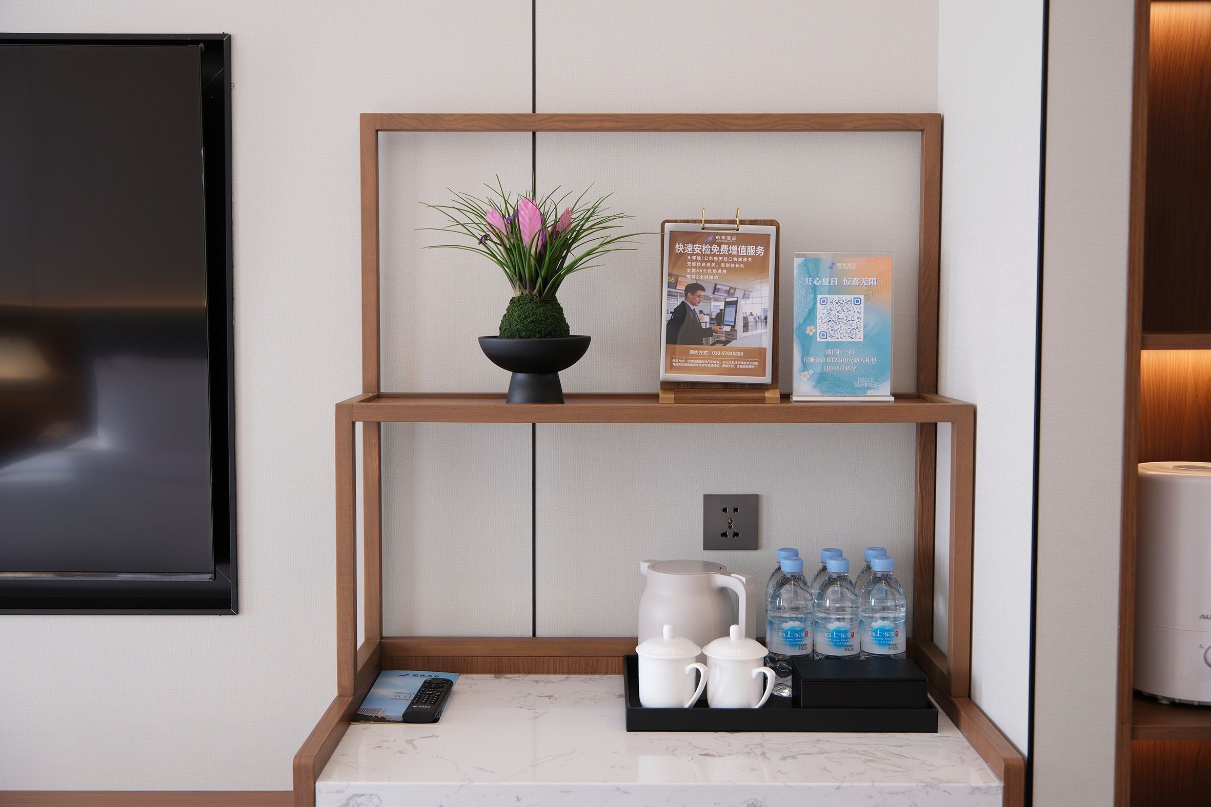 A hotel reception area with a wooden shelf displaying a potted plant, a framed informational poster, and a QR code. Below, there are two white mugs, a water bottle, and a remote control on a marble countertop.
