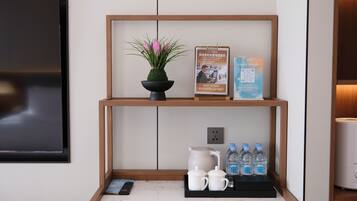 A hotel reception area with a wooden shelf displaying a potted plant, a framed informational poster, and a QR code. Below, there are two white mugs, a water bottle, and a remote control on a marble countertop.