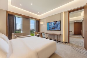Habitación de hotel con una cama grande, televisión, zona de estar con una mesa y vistas al exterior.