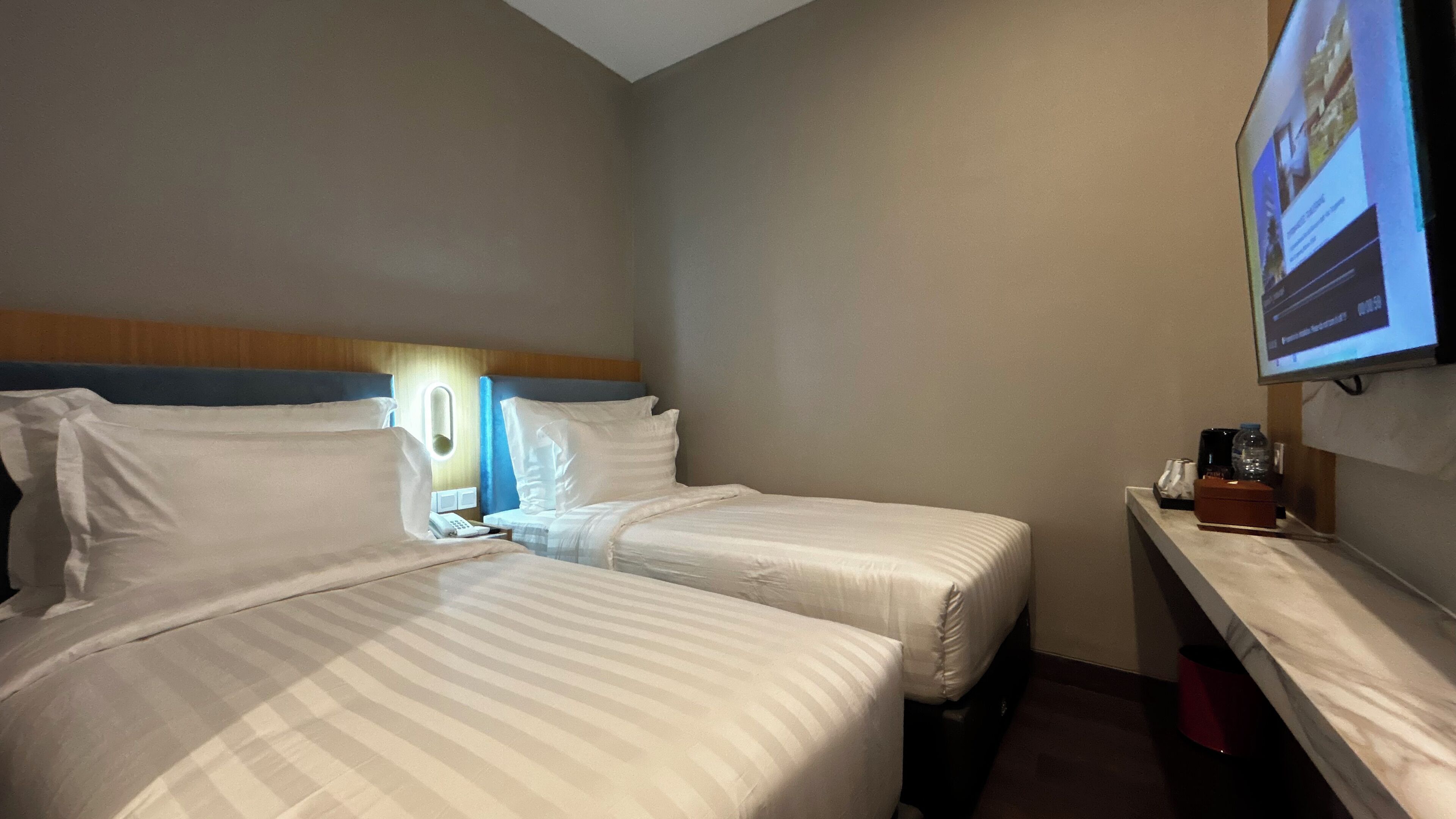 A hotel room with two beds, a bedside table, a TV, and a small desk with a computer and a bottle.