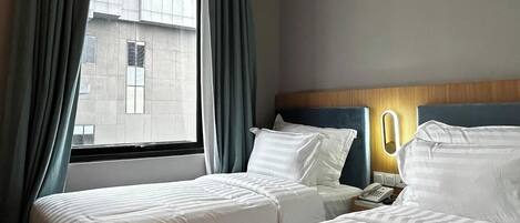 A hotel room with two beds, a window with curtains, a bedside table with a lamp, and a wall-mounted phone.