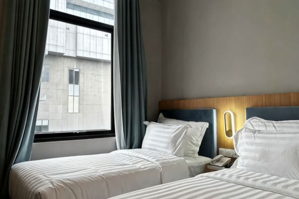 A hotel room with two beds, a window with curtains, a bedside table with a lamp, and a view of a building outside.