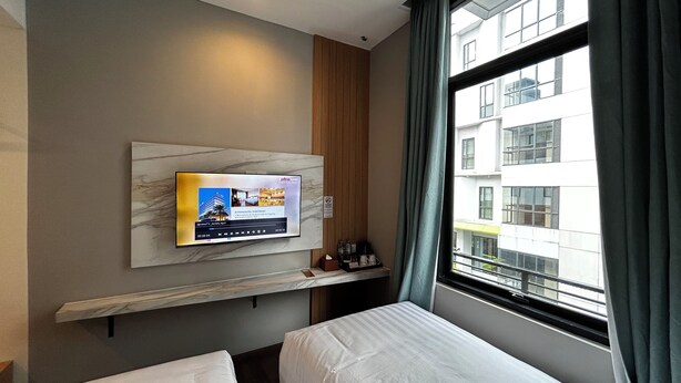 A hotel room with a television mounted on the wall displaying a hotel advertisement, a window with curtains, and a bed with a white headboard. - d'primahotel Harmoni Jakarta-Hayam Wuruk (Jakarta)