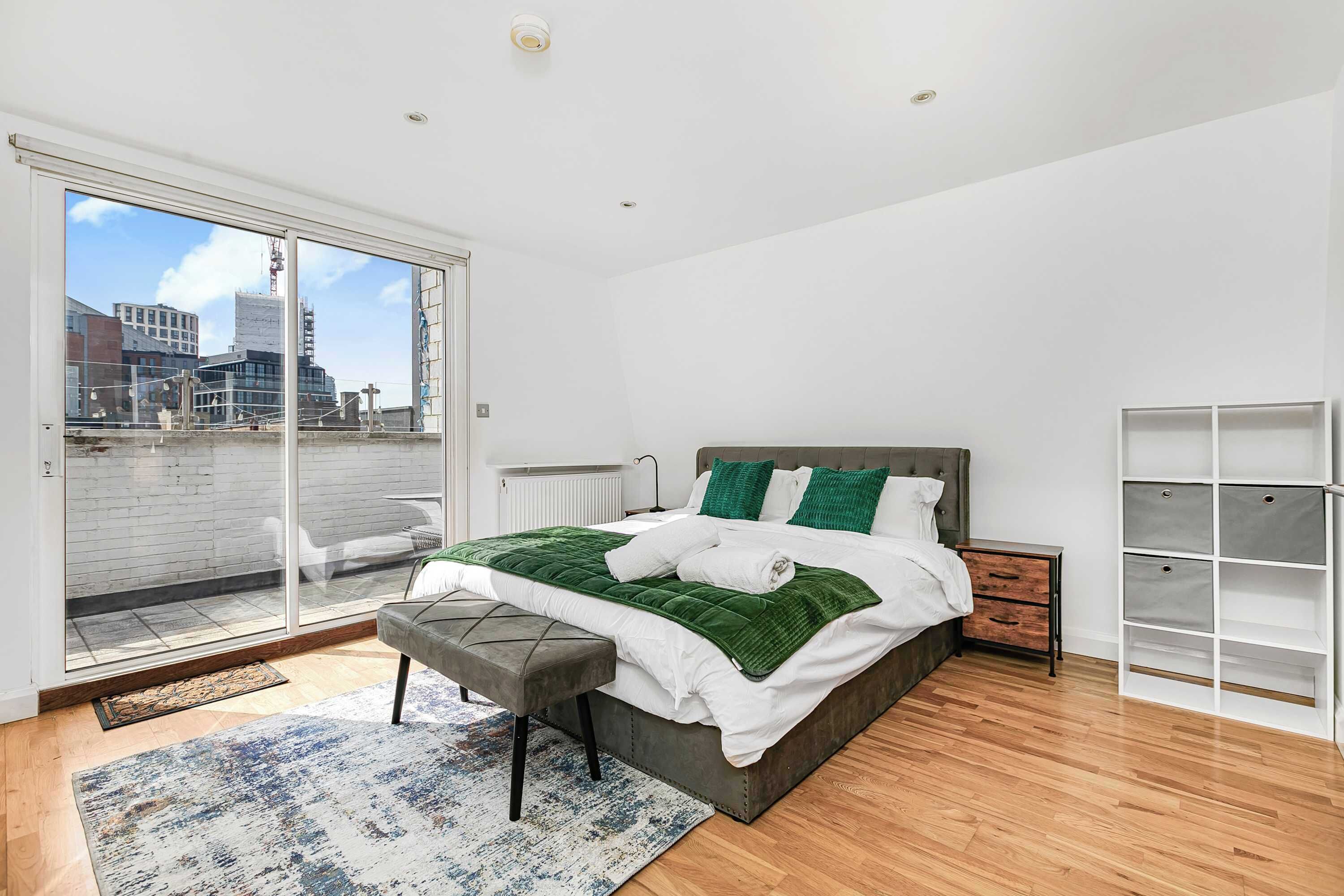 A modern bedroom with a large bed, a bench, a view of the city, and a storage unit.
