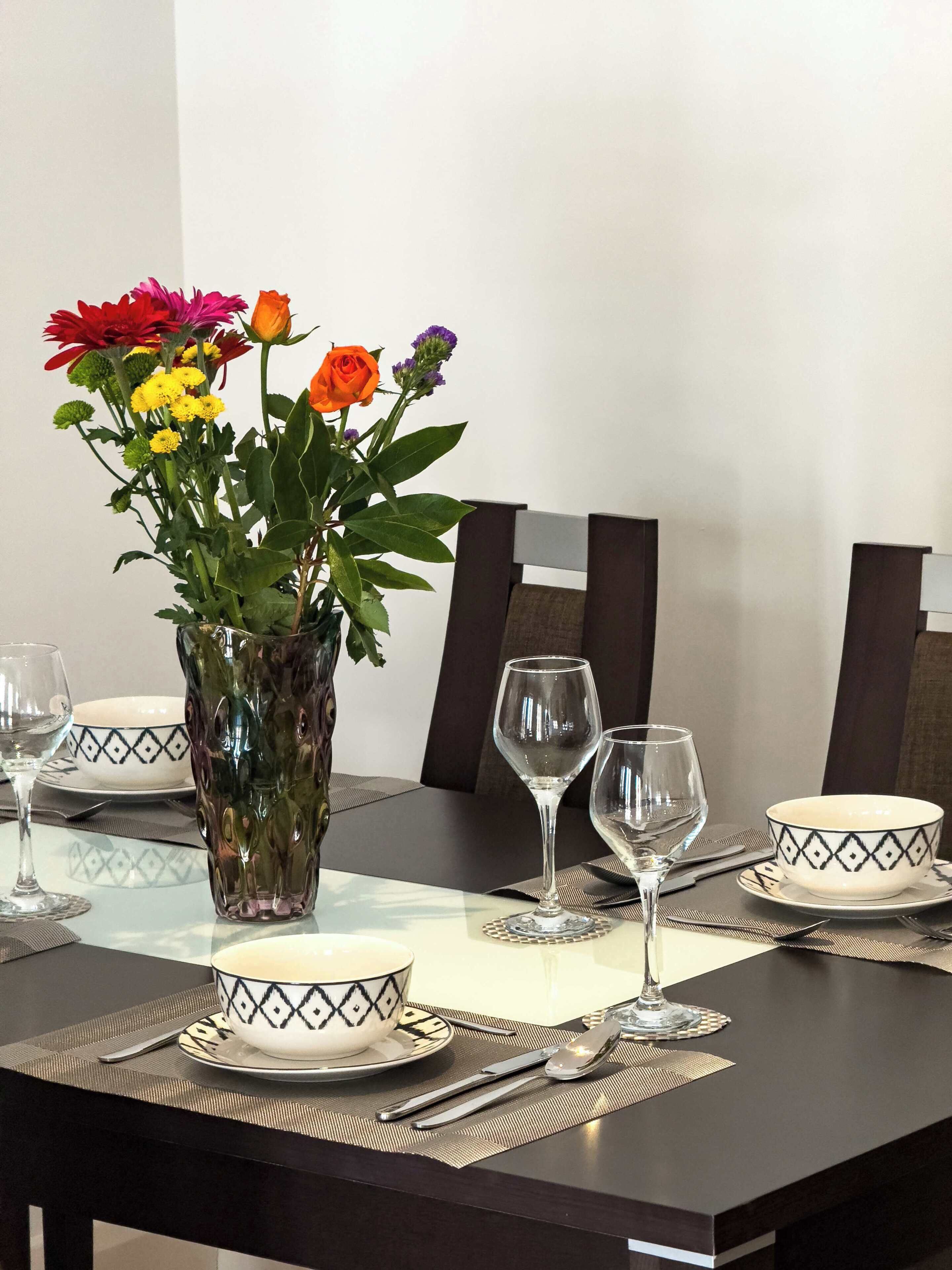 Une table à manger dressée avec des bols blancs, des verres à vin transparents et un vase rempli de fleurs.