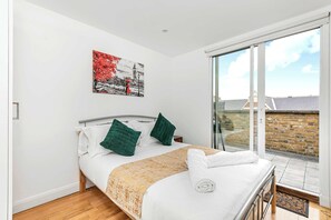 Une chambre à coucher avec un lit, des oreillers, une couverture, un tableau et une vue sur l’extérieur.