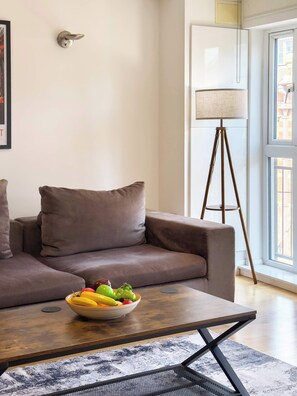 Een moderne woonkamer met een bank, een bijzettafeltje met een fruitschaal, een vloerlamp en een raam met uitzicht.