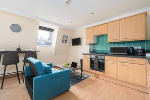 A compact kitchen with wooden cabinets, a microwave, and a stove. There is a blue sofa and a dining table with chairs.