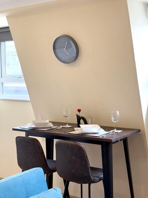 A dining area with a table set for two, a wall clock, and a window with blinds. - Cute Old Street Studio Suit Couple (Greater London)