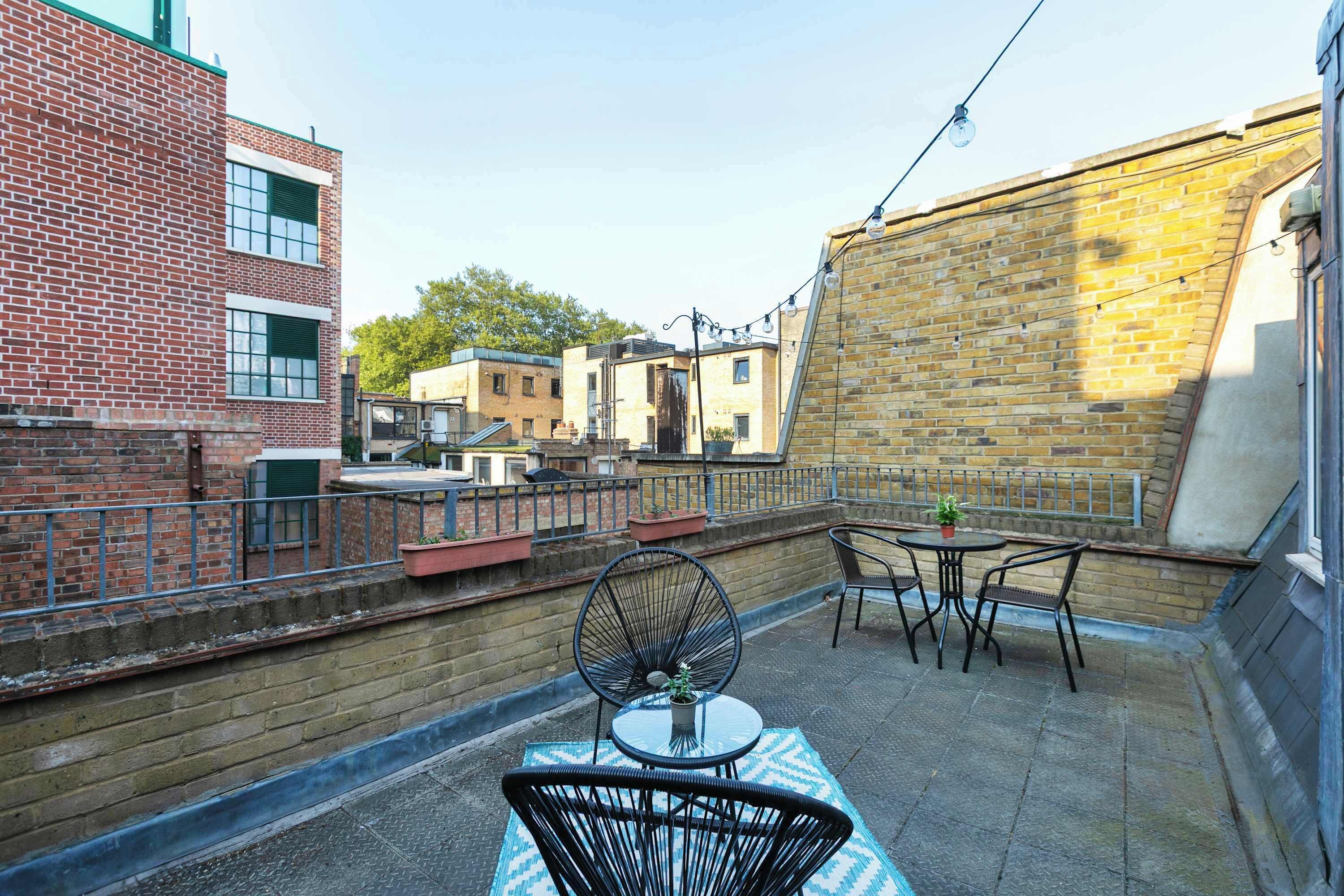 A rooftop patio with a table and chairs, a fan, and a view of neighboring buildings.